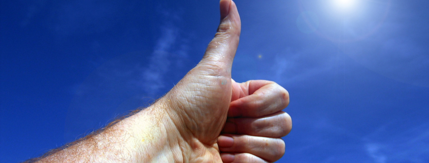 Eine linke Männerhand zeigt "Daumen rauf" vor blauem Himmel