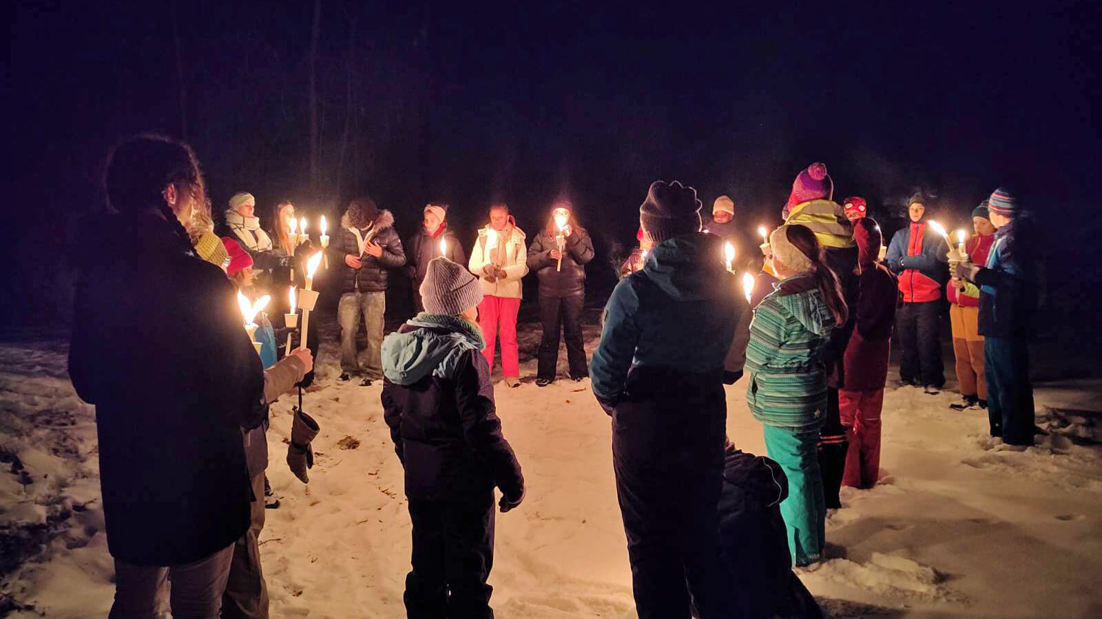 Minitrant/innen stehen nachts mit Kerzen im Kreis im Schnee